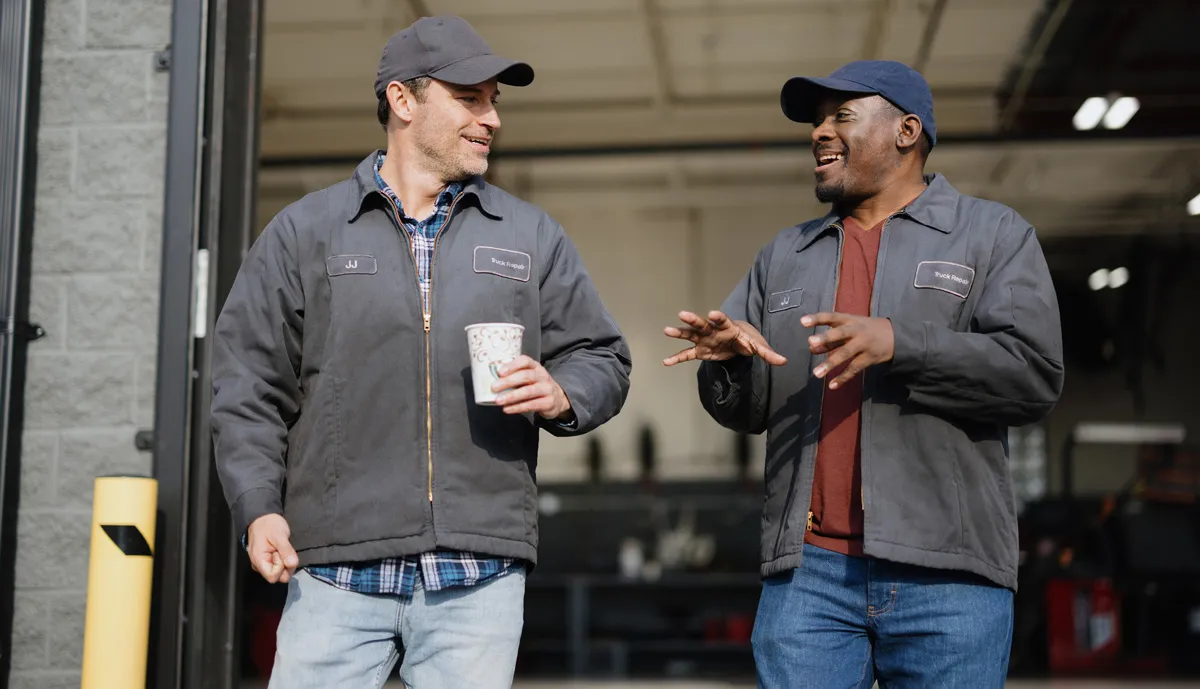 Two men in work uniforms talking and laughing outside a garage.