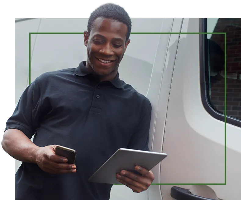 A man in a black polo uses a smartphone and tablet next to a van.