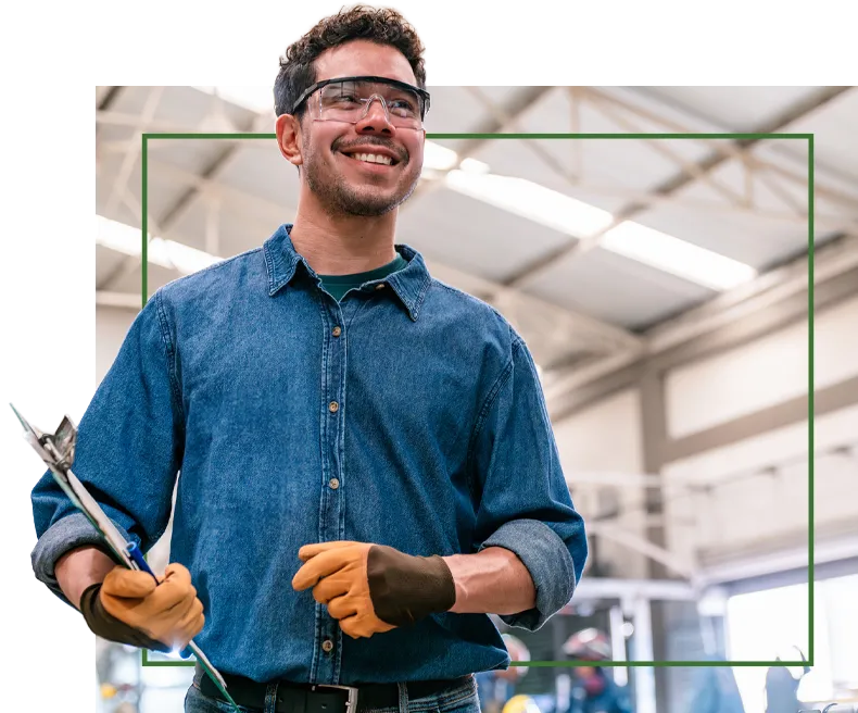 A man in safety goggles and gloves smiles while holding tools in an industrial setting.