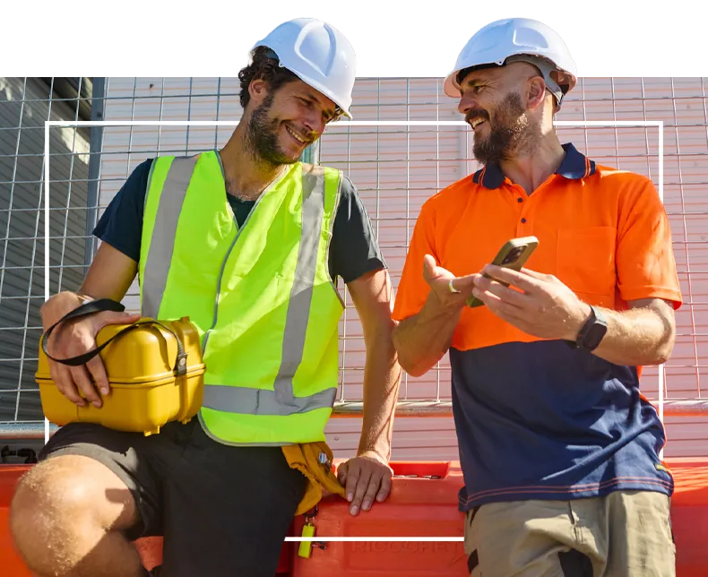 Two construction workers in safety gear talking and smiling at a construction site.