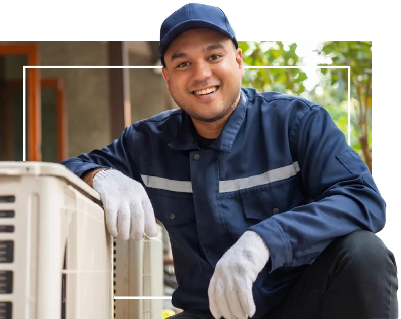 Smiling technician in blue uniform with cap and gloves kneeling beside HVAC system outdoors.