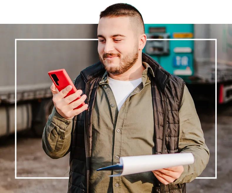 A man smiling at his phone while holding documents, standing outdoors.