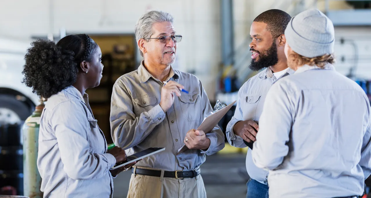 A group of factory workers in uniform having a discussion, one holds a clipboard, in an industrial setting.