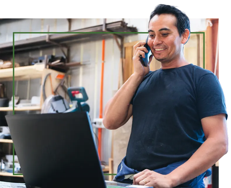 Man smiling while talking on phone and using laptop in workshop.